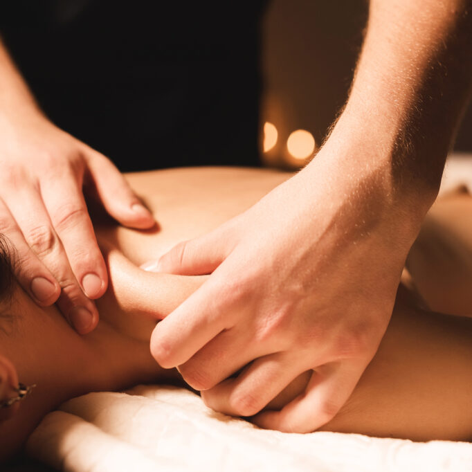 Men's hands make a therapeutic neck massage for a girl lying on a massage couch in a massage spa with dark lighting. Close-up. Dark Key.